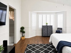Un dormitorio con cama, cómoda, televisor y ventana con contraventanas.