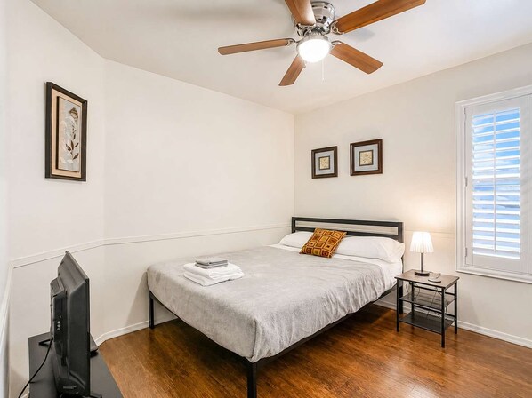 A bedroom with a bed, a ceiling fan, a television, a window with blinds, and framed artwork on the wall.