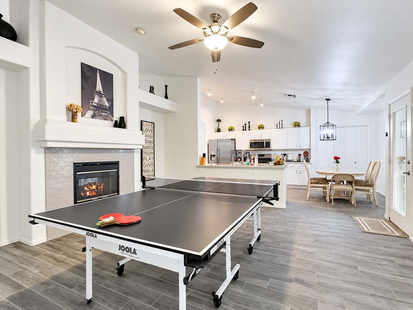 A modern living space with a ping pong table, a fireplace, a ceiling fan, and a kitchen area.