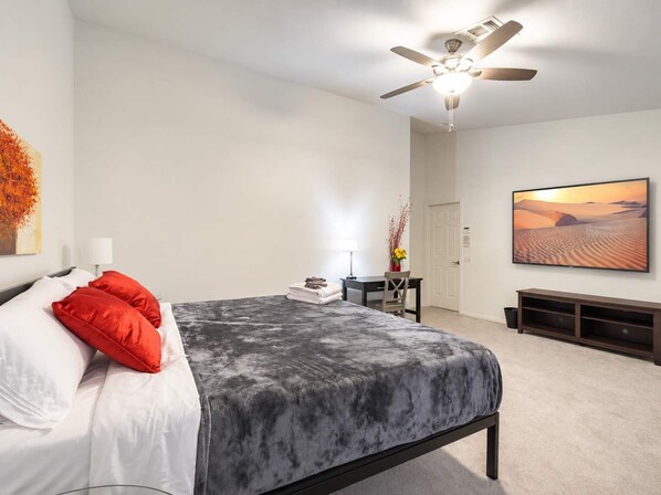A bedroom with a bed, a ceiling fan, a desk with a chair, a television, and a painting on the wall.