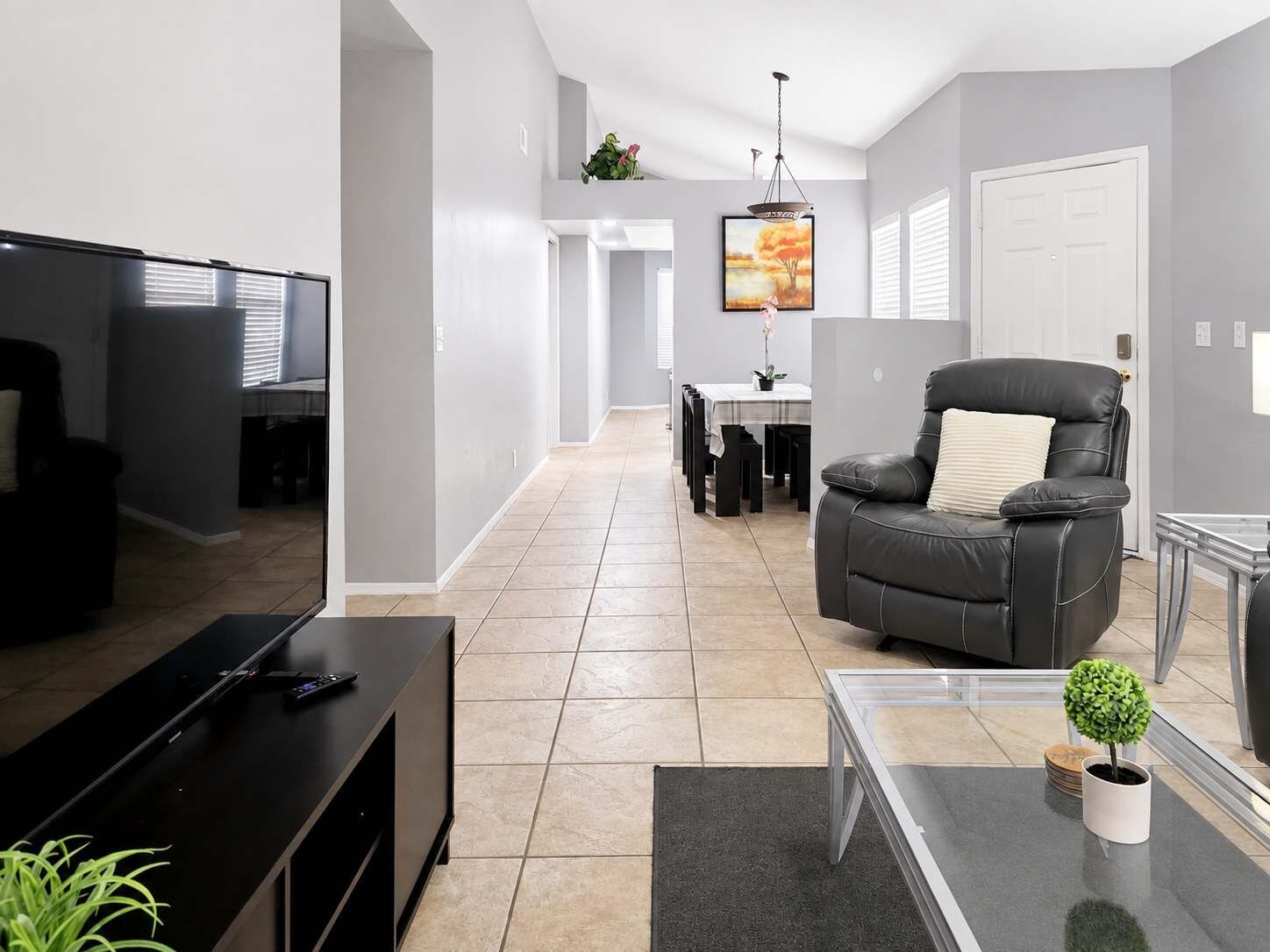 A modern living room with a flat-screen TV, a leather sofa, a glass coffee table, and a dining area in the background.