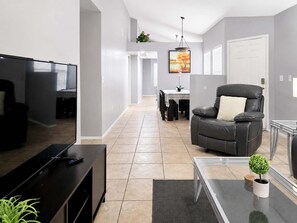 A modern living room with a flat-screen TV, a leather sofa, a glass coffee table, and a dining area in the background. - Pet-Friendly 3BR | Fenced Yard | King Bed (Chandler)