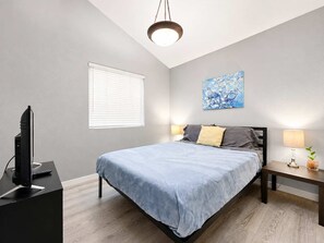 A bedroom with a bed, bedside lamps, a television on a stand, a window with blinds, and a painting on the wall. - Pet-Friendly 3BR | Fenced Yard | King Bed (Chandler)