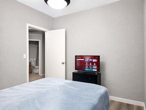 A bedroom with a bed, a television on a stand, a door leading to a bathroom, and a ceiling light. - Pet-Friendly 3BR | Fenced Yard | King Bed (Chandler)