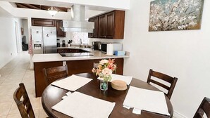 A dining area with a round table, four chairs, a ceiling fan, and a kitchen in the background.