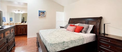 A bedroom with a wooden bed, a nightstand, a dresser, a ceiling fan, and a bathroom visible in the background.