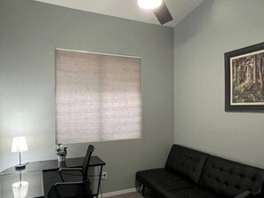 A room with a dark sofa, a small table with a lamp, and a window with blinds.