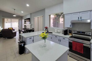 A modern kitchen with white cabinets, a central island with a vase of flowers, a built-in oven, and a dining area with a brown sofa and a table. - Pet-Friendly 3BR | Private Pool | Game Room (Chandler)