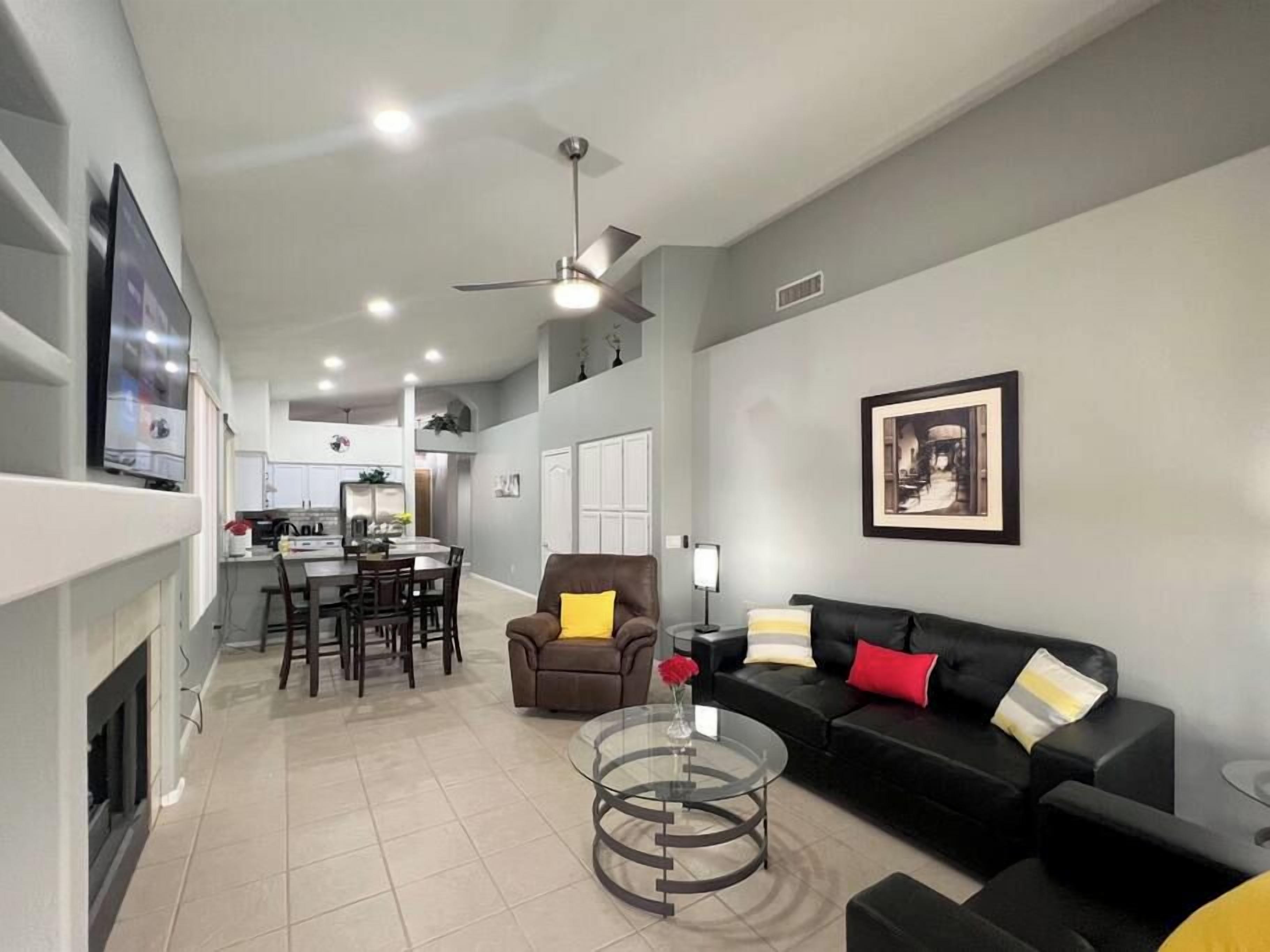 A modern living room with a black sofa, a glass coffee table, and a dining area with a kitchen in the background.