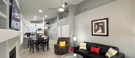A modern living room with a black sofa, a glass coffee table, and a dining area with a kitchen in the background.