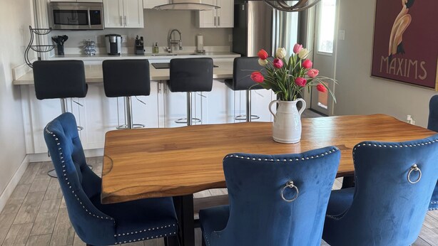 Un coin repas avec une table en bois, des chaises à l’assise rembourrée de couleur bleue, un lustre et, au fond, une cuisine.