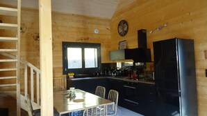 A wooden cabin interior with a kitchen area, including a dining table with chairs, a refrigerator, and a window.