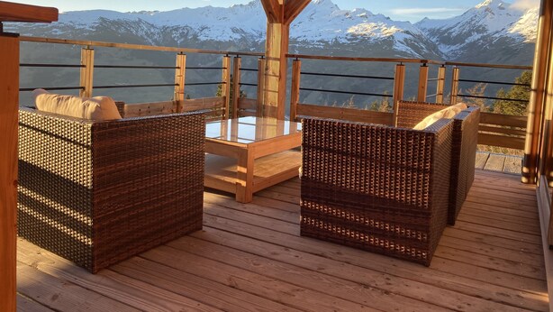Une terrasse en bois, agrémentée de meubles en osier et d’une table en verre, offrant une vue sur des montagnes enneigées.
