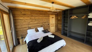 Une chambre avec un plafond en bois, un lit avec une literie blanche, un luminaire suspendu et un dressing où les vêtements sont accrochés.