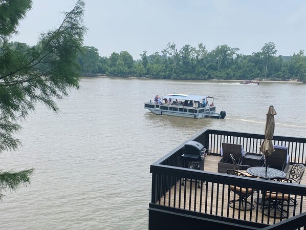 Vista de un río desde una terraza con mobiliario exterior y un barco al fondo.