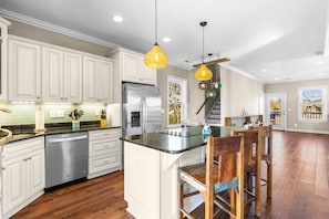 Una cocina con armarios blancos, un refrigerador de acero inoxidable, una mesa de comedor de madera con sillas y luces colgantes.