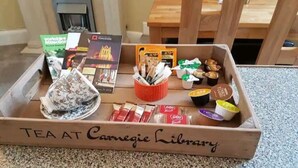A wooden tray with a tea service, books, and snacks labeled "TEA AT Carnegie Library".
