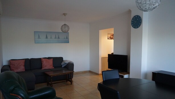 A living room with a sofa, coffee table, and chairs, a dining table, a TV, and a painting on the wall. - Quiet F4 apartment 5 minutes from Olhão town center, Algarve  (QUELFES)