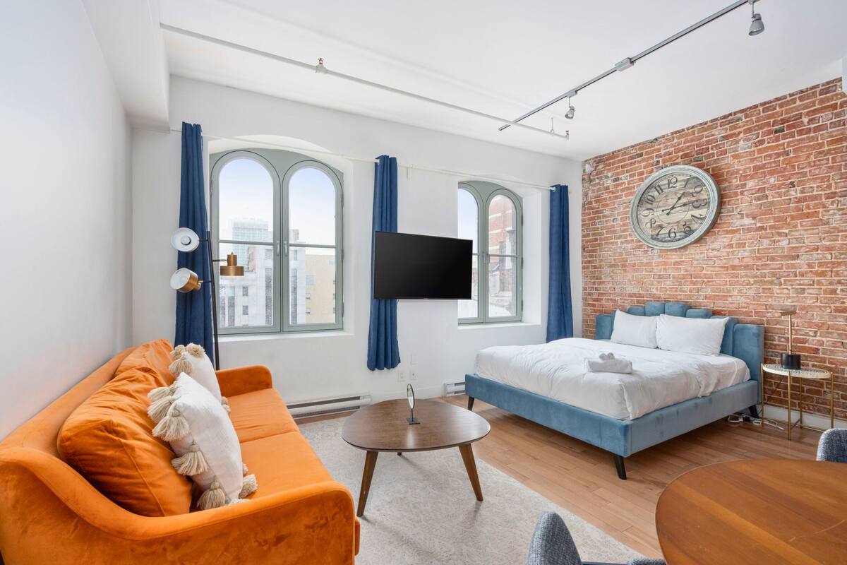 A modern hotel room with a large bed, a sofa, a TV, and a brick wall.