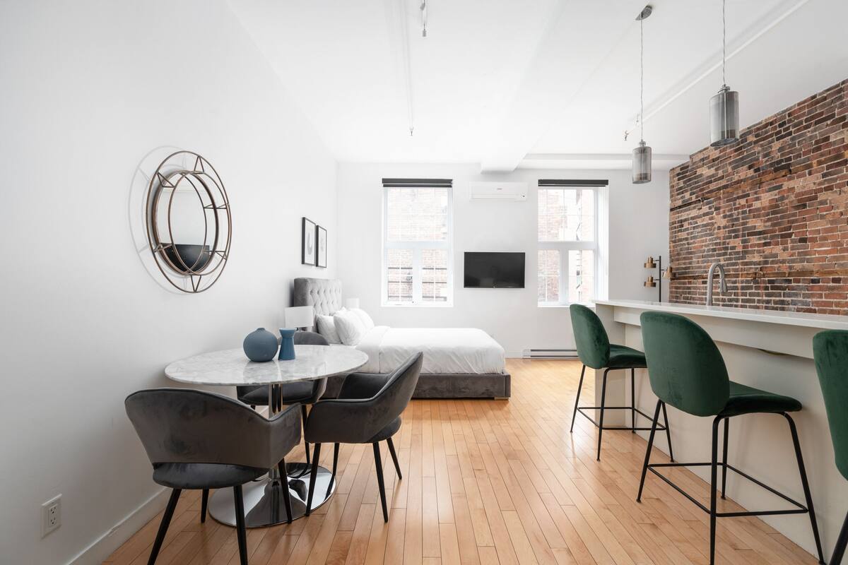 A modern room with a bed, a dining table with chairs, a kitchen area with bar stools, and a large window providing natural light.