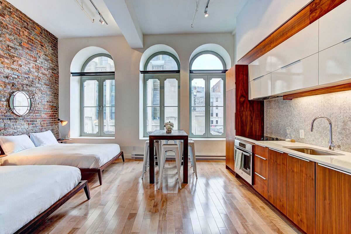A modern kitchen with wooden cabinets, a sink, and a dining area with a table and chairs. There are large windows with arched tops, and a brick wall on one side.