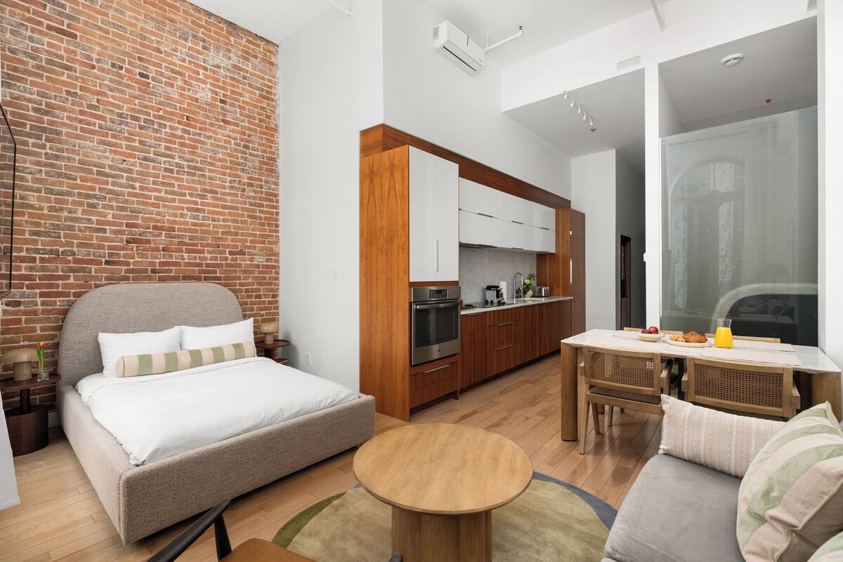 A modern room with a bed, a dining table, a kitchen area, and a large window.