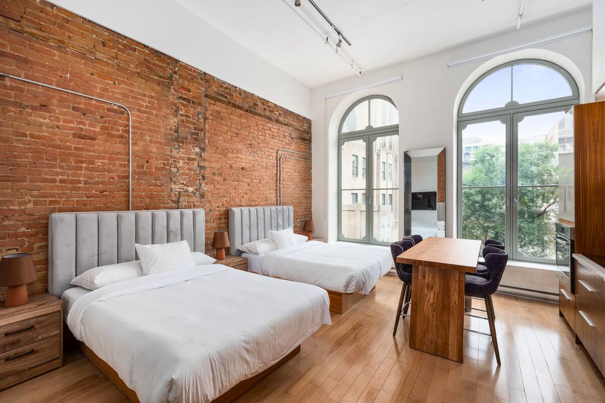 A modern hotel room with two beds, a dining table, and large windows offering a city view.