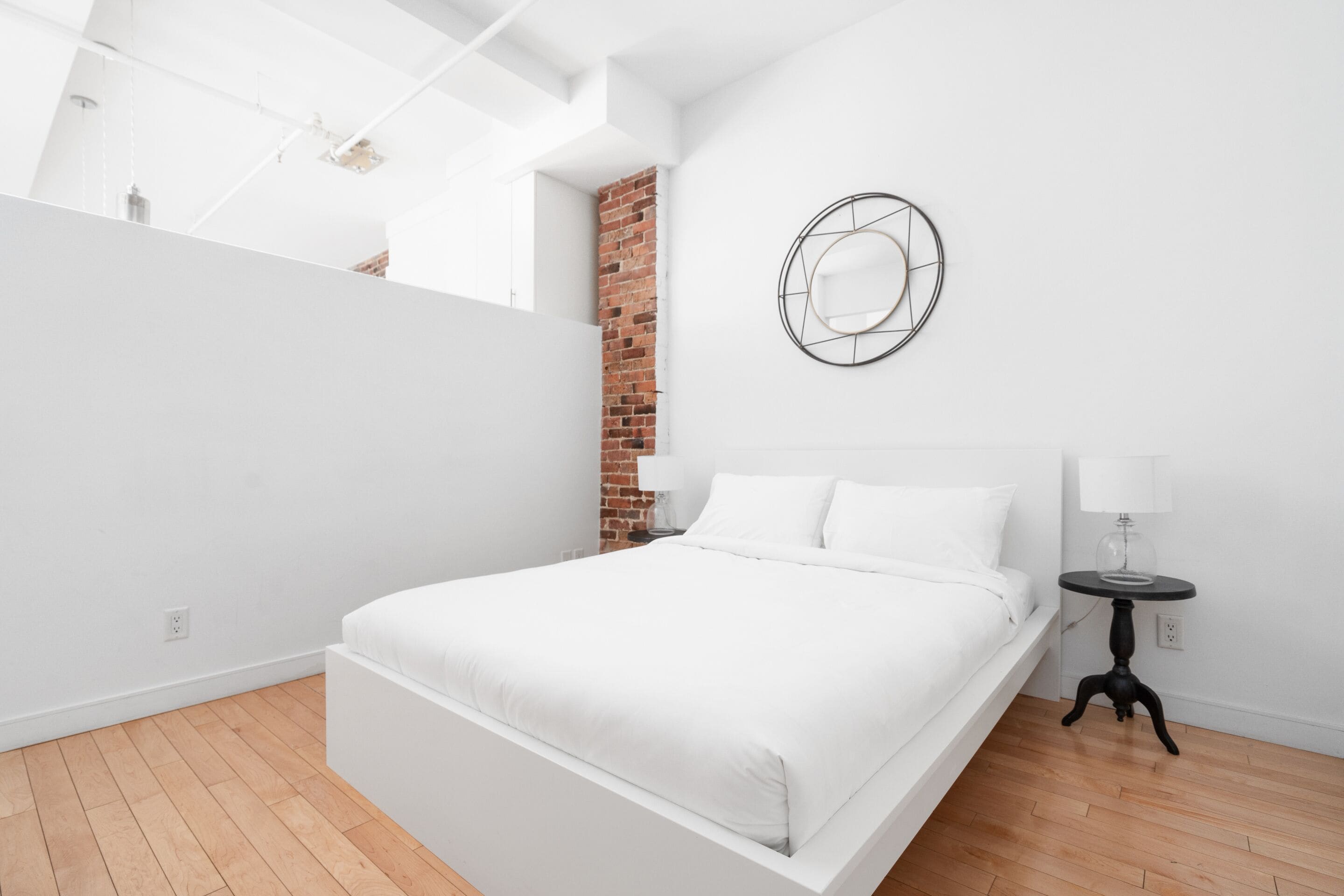 A modern bedroom with a white bed, a brick wall, and a round mirror on the wall.