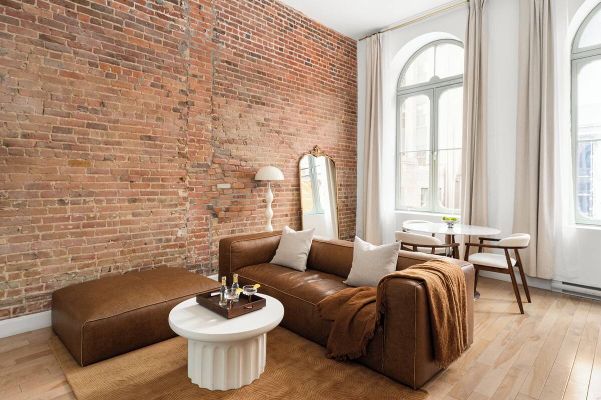 A room with a brick wall, leather sofa, round coffee table, and a dining area with a table and chairs.