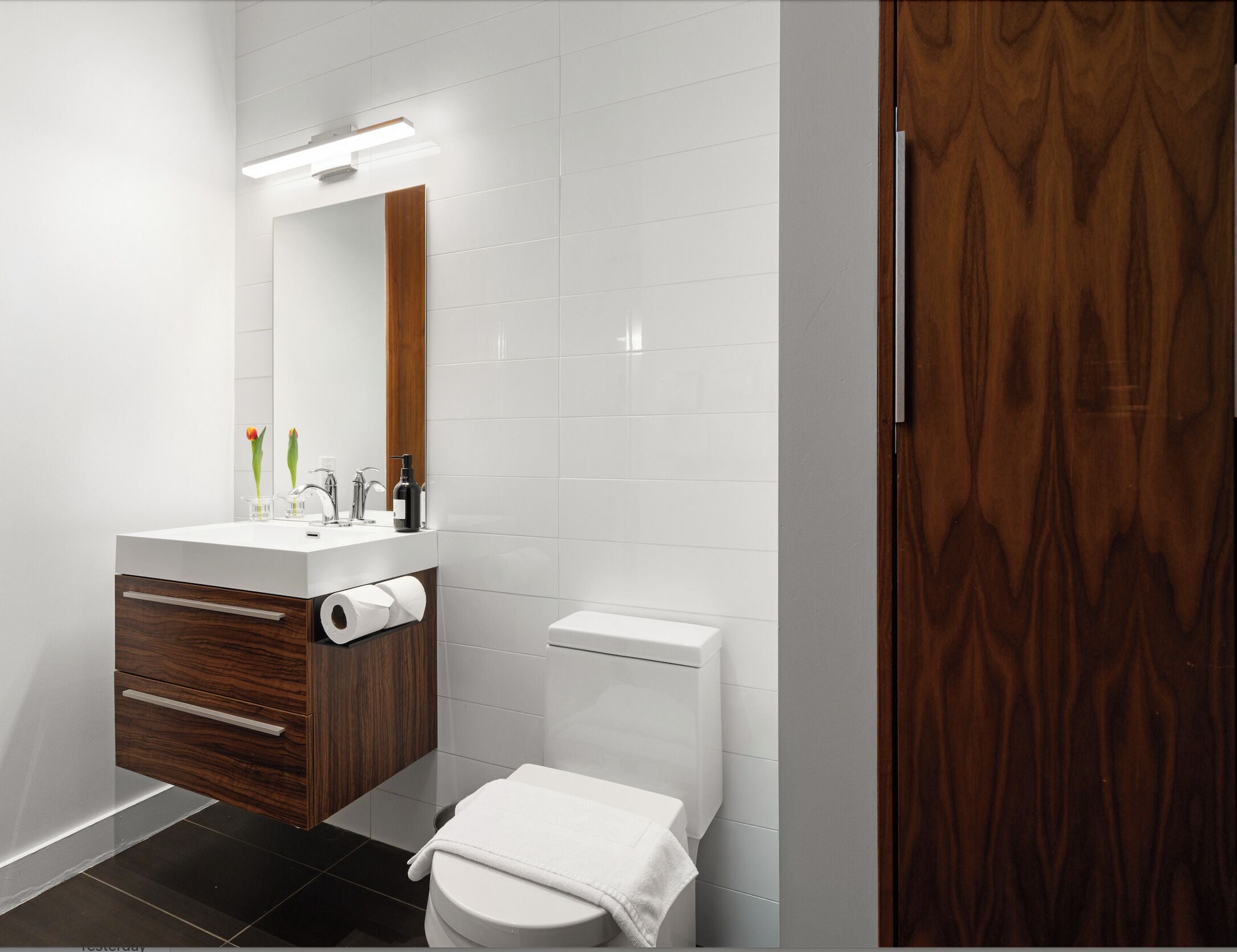 A modern bathroom with a white vanity, a mirror, a toilet, and a wooden door.