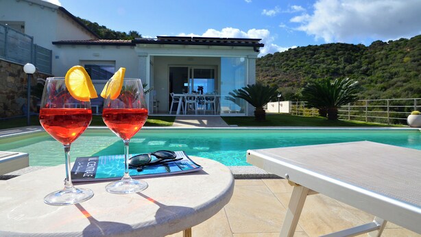 A poolside table with two glasses of red wine, a pair of sunglasses, and a magazine.