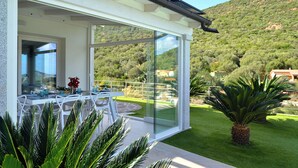A modern outdoor dining area with a glass enclosure, white furniture, and a view of a lush hillside.