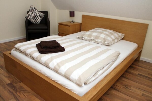 Una cama bien hecha con ropa de cama a rayas, un cabecero de madera, una mesita de noche con una lámpara y una silla negra con un cojín estampado.