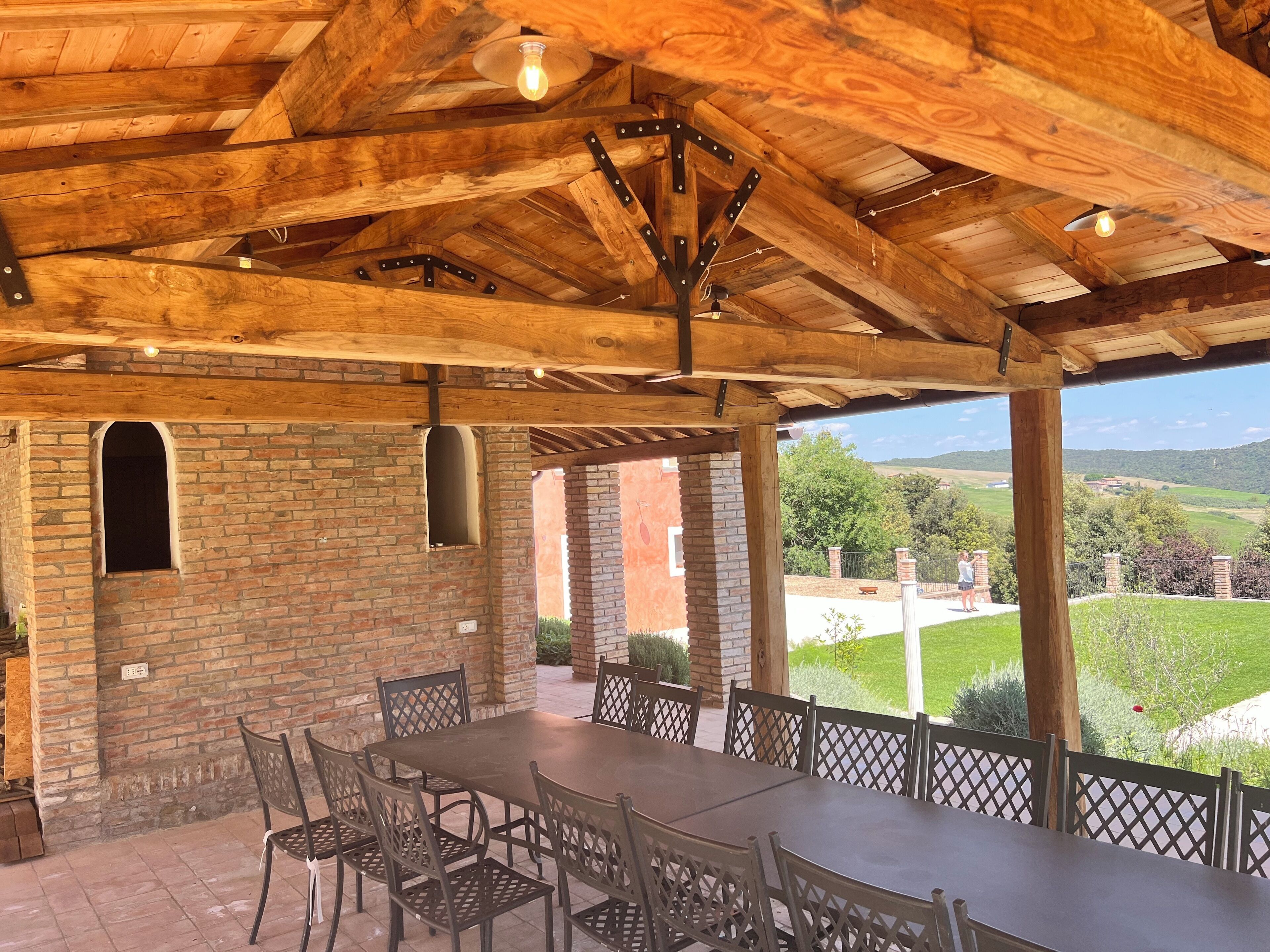 Un espacio de comedor al aire libre techado, con pared de ladrillo, vigas de madera y vistas al campo.