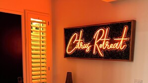 Um corredor com um letreiro de néon que diz "Citrus Retreat" na parede, uma porta com persianas, uma mesinha de apoio com um vaso e um vaso decorativo no chão.