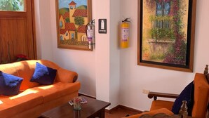 A living room with an orange sofa, a wooden chair, a small table, and two paintings on the wall. - Hotel Sol De La Villa (Villa de Leyva)
