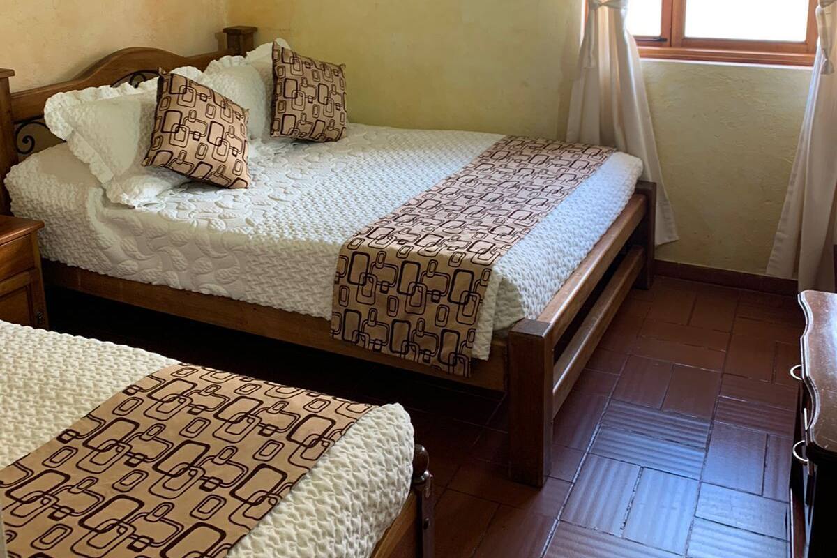 Quarto com duas camas de madeira, cada uma com roupa de cama estampada e almofadas. Há uma janela com cortinas, e o chão é de azulejos.