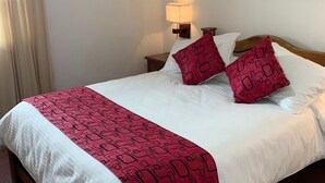 A hotel room with a bed, a nightstand with a lamp, a painting on the wall, and a window with curtains. - Hotel Sol De La Villa (Villa de Leyva)