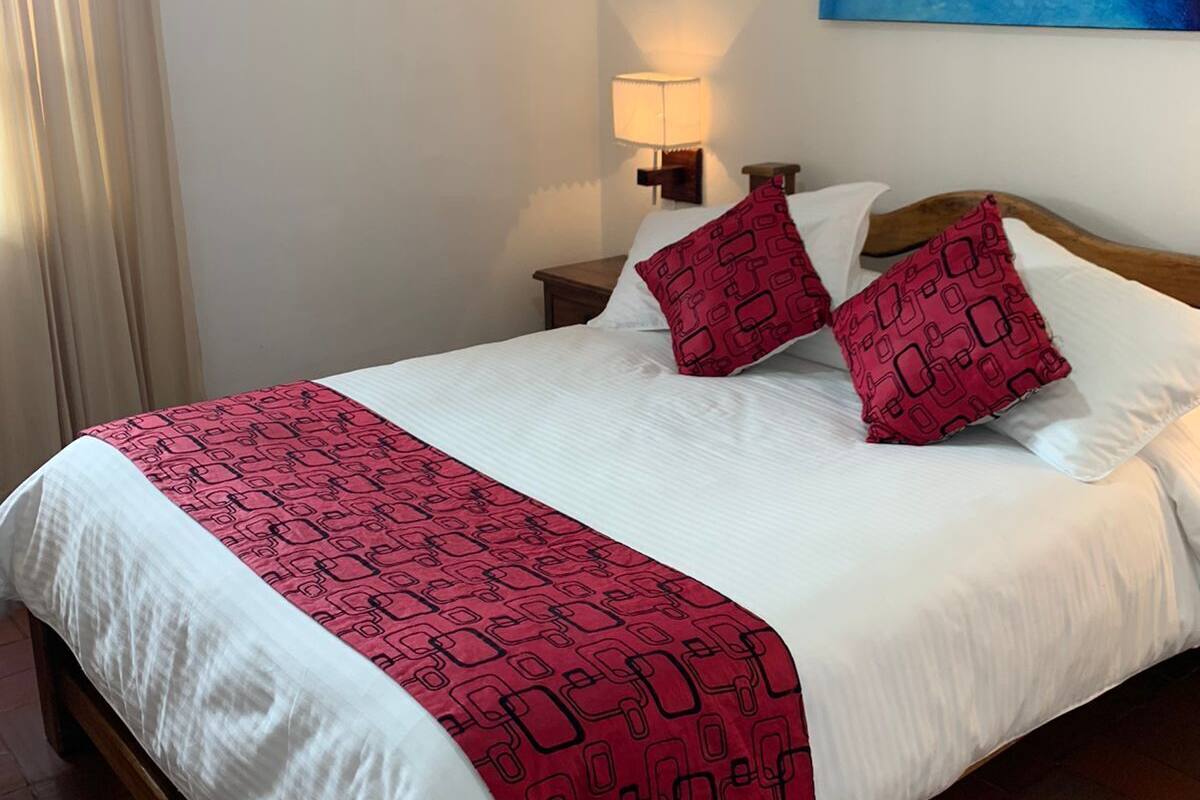 Quarto de hotel com uma cama, uma mesinha de cabeceira com abajur, um quadro na parede e uma janela com cortinas.
