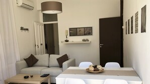 Ein kompakter Wohnbereich mit Sofa, Esstisch mit Stühlen, einem kleinen Couchtisch, Klimaanlage, Pendelleuchte, einer Tür und einer Wandregal mit Dekoration.