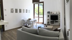 Ein modernes Wohnzimmer mit einem grauen Sofa, einem Esstisch mit Stühlen, einem Fernseher auf einem weißen Regal und Kunstwerken an der Wand.