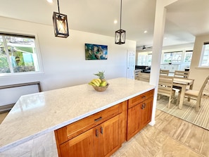 A kitchen with a marble countertop, wooden cabinets, a fruit bowl, pendant lights, and a painting on the wall.