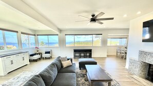 Amplia sala de estar con un sofá seccional gris grande, una mesita de madera y una chimenea. Varias ventanas ofrecen vistas al océano.