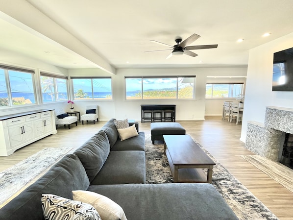 A spacious living room with a large gray sectional sofa, a small wooden table, and a fireplace. There are multiple windows offering a view of the ocean.