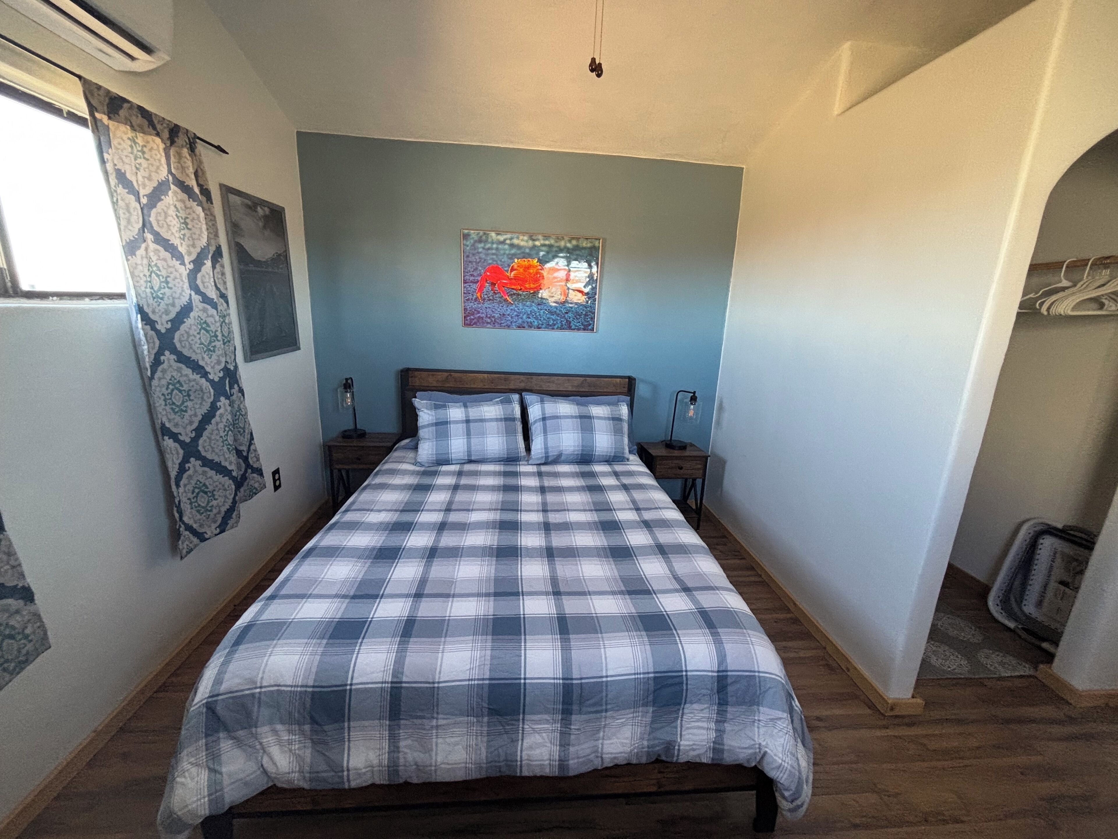A bedroom with a bed, two bedside tables, a wall-mounted artwork, a window with patterned curtains, and a radiator.