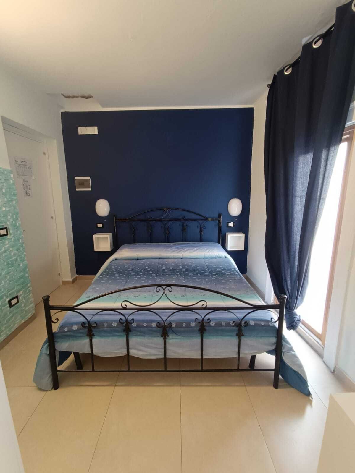 A bedroom with a bed, a nightstand, a wall-mounted lamp, and a window with curtains.