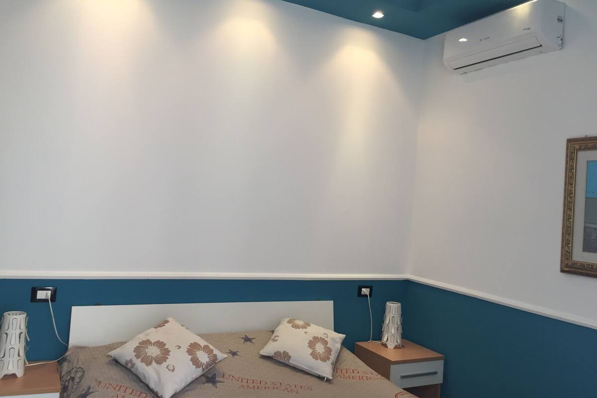 A bedroom with a bed, two bedside tables, an air conditioner, and a wall-mounted picture frame.