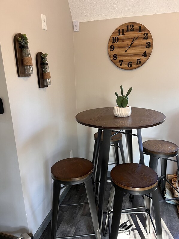A dining area with a round wooden table, four stools, and a wall clock. - Rustic Boondock - Peaceful tiny home on 143 farm acres… down a scenic dirt road! (Eureka Springs)