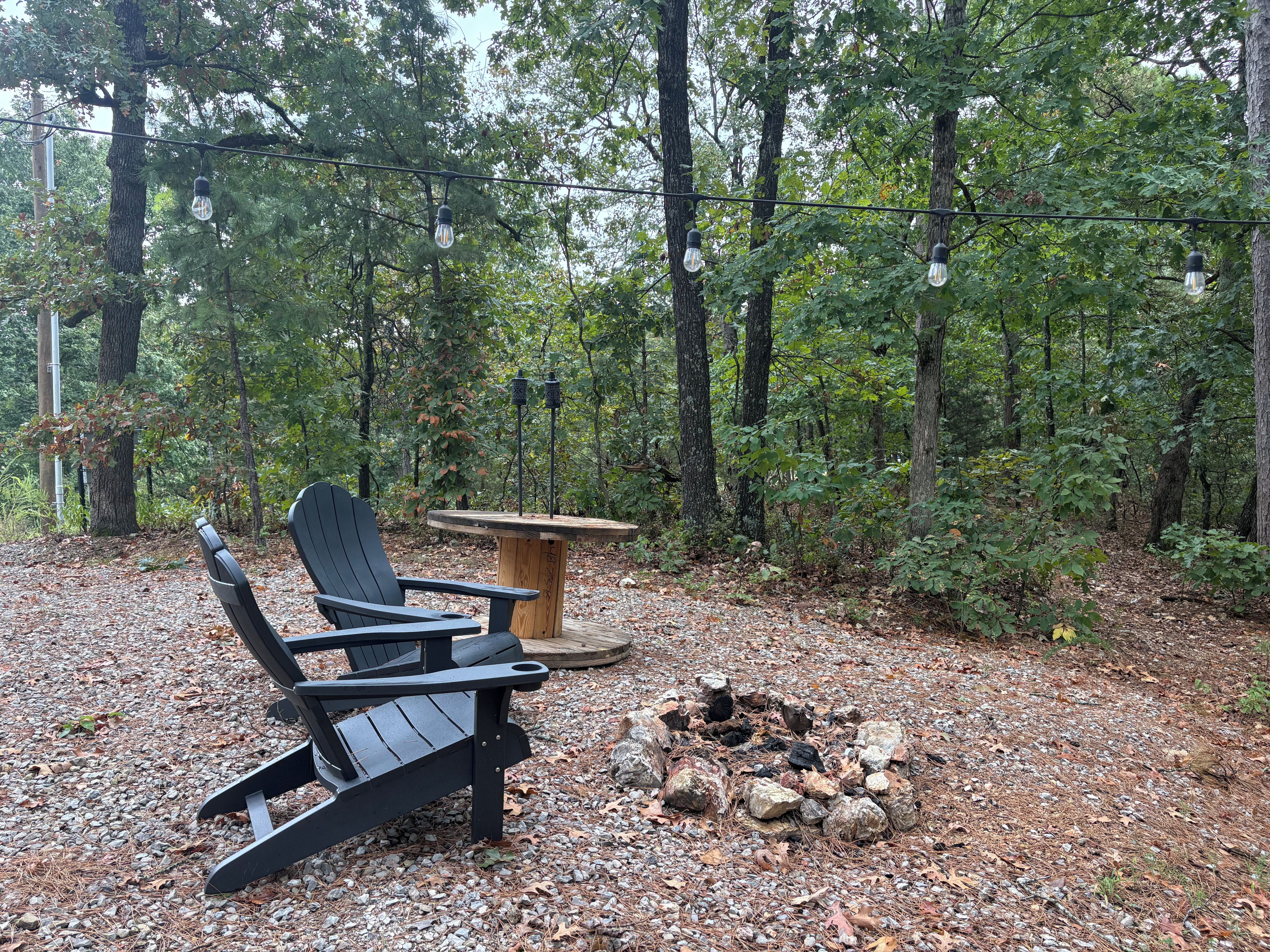 A forested area with a wooden table and two Adirondack chairs.