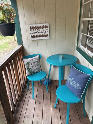 Una terraza de madera con dos sillas turquesa y una mesita pequeña, una maceta colgante y un cartel con una escena de bosque y el texto 'Let's go exploring'.